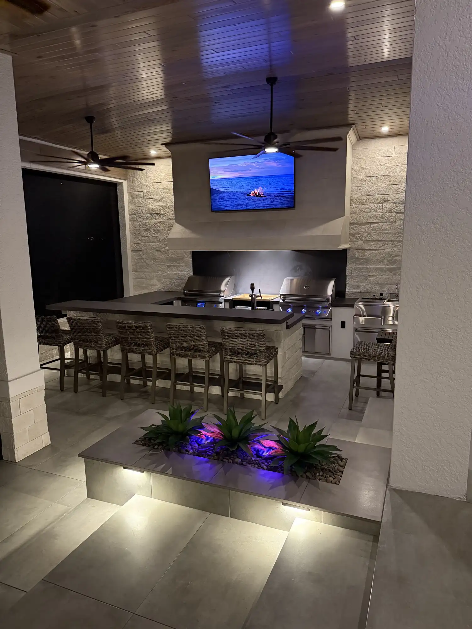 The finished outdoor living space lit at night with bar stools and TV wall